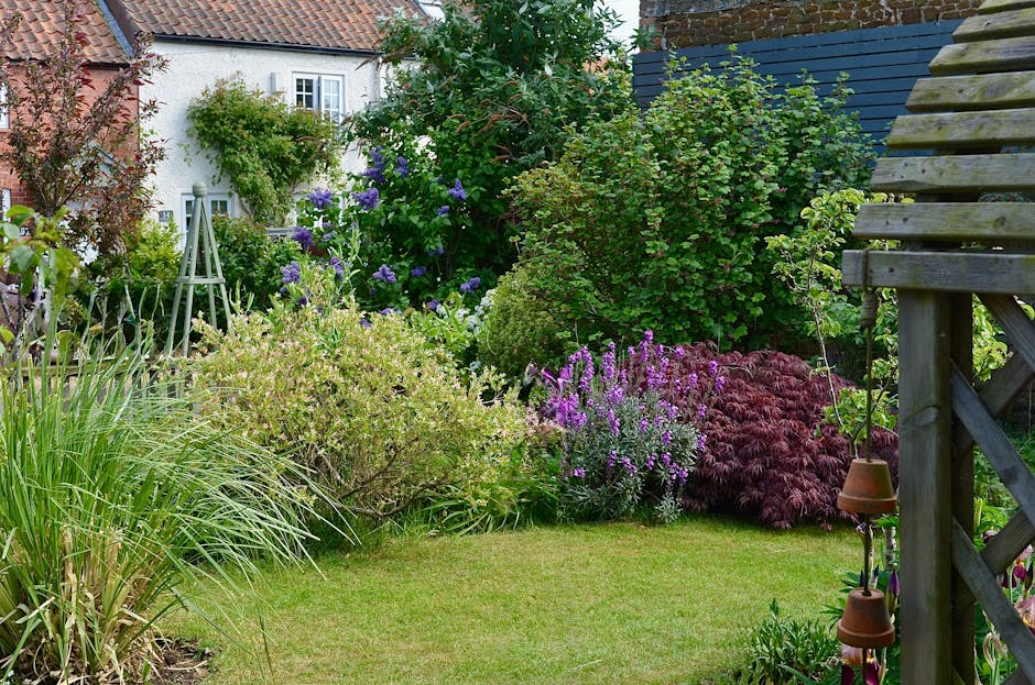 A small, well-maintained garden in Cranford featuring a neatly trimmed lawn bordered by lush, flowering shrubs and plants. In the foreground, there is a patch of bright green grass with a variety of p